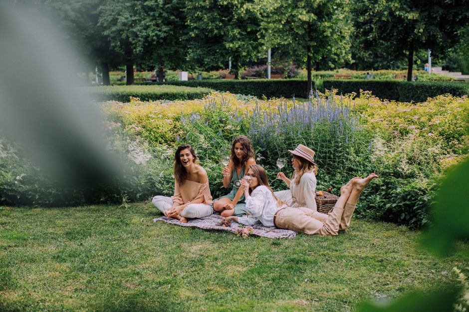 A group of four women enjoying a picnic on lush green grass in a park with vibrant flower beds and tall trees in the background. They are seated on a blanket, surrounded by a variety of fresh flowers
