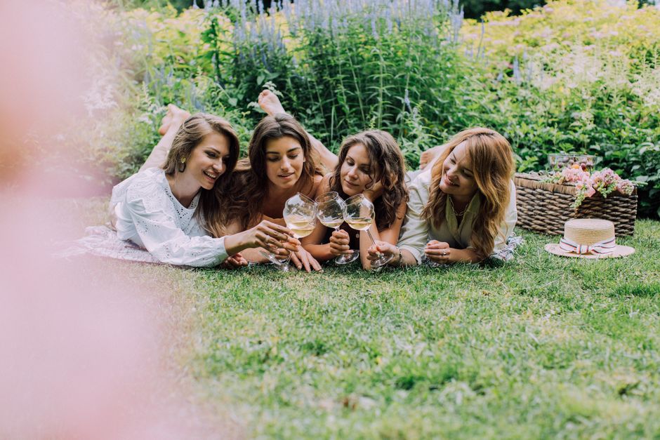 Four women lying on their stomachs on a lush green lawn during a picnic in a garden surrounded by tall purple and green plants, holding and clinking glasses of pale white wine or champagne, all smilin
