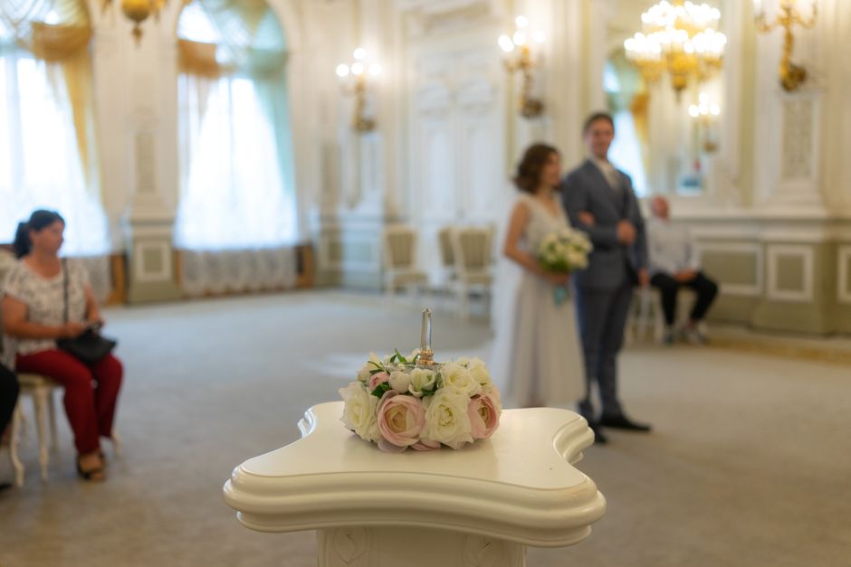 A close-up of a floral arrangement featuring a small bouquet of cream and pale pink roses, with delicate greenery, placed on a white ornate pedestal in a bright, elegant room. In the background, a bri