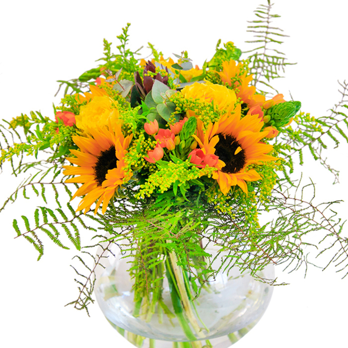 Brimming with the joyful palette of Watford's best-loved spaces, this elaborate bouquet celebrates both the artistry of floristry and the region's natural abundance. Majestic sunflowers, their bold yellow petals flaring out from rich, brown centres, take pride of place-a fitting homage to the wildflowers seen along Cassiobury Park's sunlit meadows. Enveloping these are elegant yellow garden roses, petals arranged so precisely they seem to glow, their spiralled forms bringing a classic sophistication worthy of a Watford Town Hall celebration. Glossy red Hypericum berries are sprinkled through the bouquet, shining against backdrop of layered green foliage and whispering of orchard walks along the local allotments. Wispy stems of solidago and goldenrod, soft feathery ferns, and delicate bluish eucalyptus leaves fill spaces with texture and hint at the gentle stir of a summer wind. All of this abundance springs from a crystal-clear, round glass vase, where submerged stems interlace in the water-a simple nod to modern minimalism set against the arrangement's rich textures. The scent that rises is light yet uplifting, weaving together memories of gardens, picnics, and long conversations by the pond. This bouquet, crafted by an experienced local florist, is ideal for milestone celebrations, heartfelt congratulations, or inviting the fresh cheer of Watford's scenery into your home.
